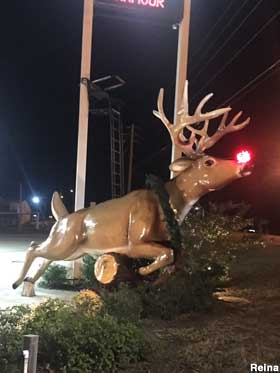 Fiberglass, full-color statue of a deer with antlers jumping over a log. Nighttime photo, and the deer has a glowing red nose.