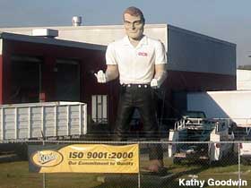 Outdoor full-color statue of a giant man behind a fence wearing a white shirt and black pants. His upturned hands are wearing white gloves.