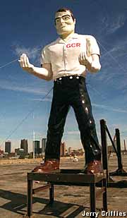 Outdoor full-color statue of a giant man wearing a white shirt and black pants.