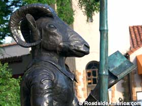 Outdoor bronze statue of a ram's head on a human body holding a book.