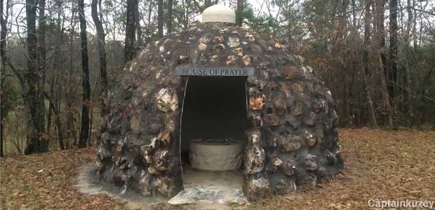 Small, dome-shaped building made of dark rocks has an open doorway, is surrounded by trees.