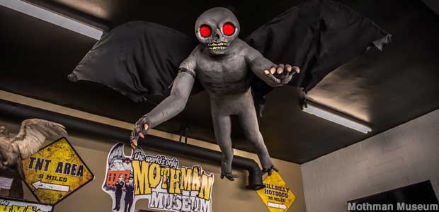 Mothman costume hangs from the museum ceiling, with a gray body, large black wings, hands with claws, a skull-like head, and glowing red eyes. He looks more like a man than a moth.