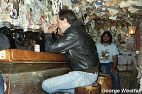 Two men sit at a bar. The walls and ceiling are covered with tacked on dollar bills and underwear.