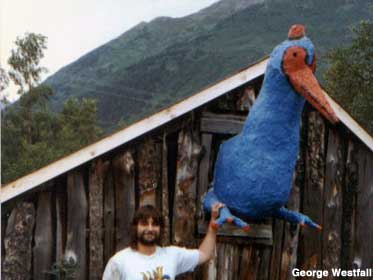 Bearded young man rests hand on leg of large blue bird sculpture poking out of a wood building.