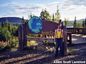 Man stands next to outdoor sign marking the Arctic Circle.
