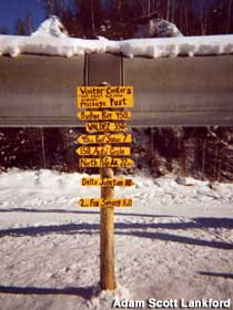 Yellow sign next to the Alaska Pipeline gives distances to various pipeline towns.