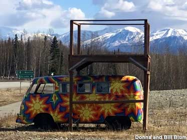 Old 1960s minibus painted in psychedelic colors sits outside beneath a wooden shelter.