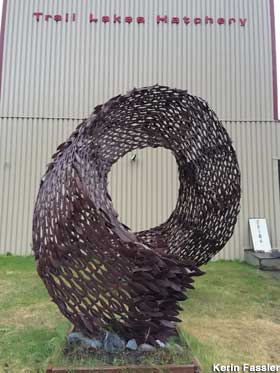 Metal sculpture of a swirling school of fish stands outside a fish hatchery building.