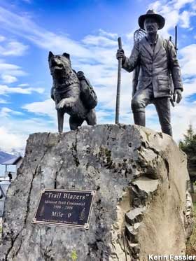 Bronze statue of a dog and a man with snowshoes and a walking stick atop an outdoor rock.