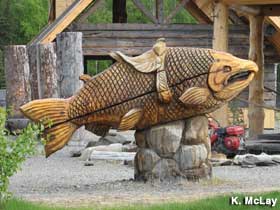 Large, outdoor, carved wooden fish with a human-size saddle on its back.