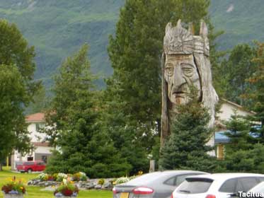 Large wooden carving of a Native American head looks out over trees and parked cars.