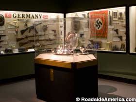 Silver tea service inside a free-standing glass case with Nazi relics displayed in wall cases behind it.