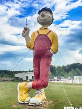 Cartoony full-color giant boy statue wears purple overalls and big sneakers, also has a big nose and smile.