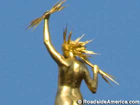 Rear view of gold statue of naked woman with lightning bolt hair and lighting bolts grasped in both hands.