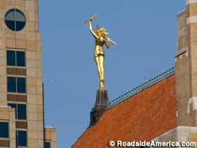 Rear view of gold statue of naked woman with lightning bolt hair and lighting bolts grasped in both hands, perched atop a spire.