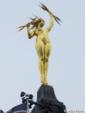 Gold statue of naked woman with lightning bolt hair and lighting bolts grasped in both hands, perched atop a spire.