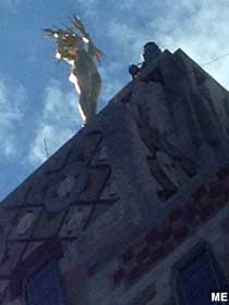 View from below of gold statue of naked woman with lightning bolt hair and lighting bolts grasped in both hands, perched atop a spire.