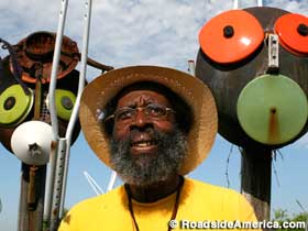 Bearded, African-American man wearing hat and yellow t-shirt stands in front of outdoor folk art African sculptures.