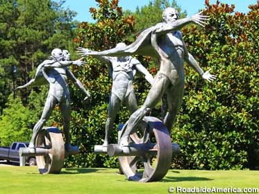 Outdoor bronze statue of three men wearing capes and riding unicycles.