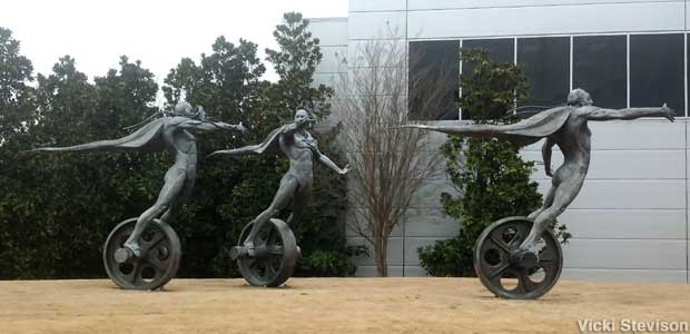 Outdoor bronze statue of three men wearing capes and riding unicycles outside a building.