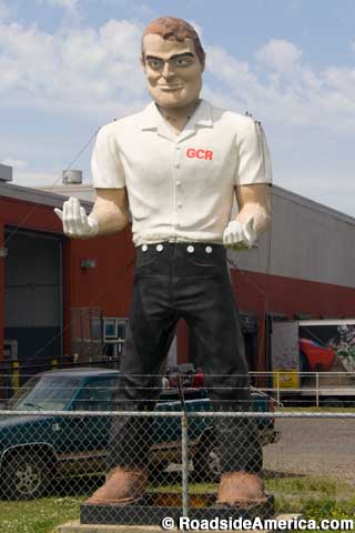 Outdoor full-color statue of a man behind a fence wearing a white shirt and black pants. His upturned hands are wearing white gloves.