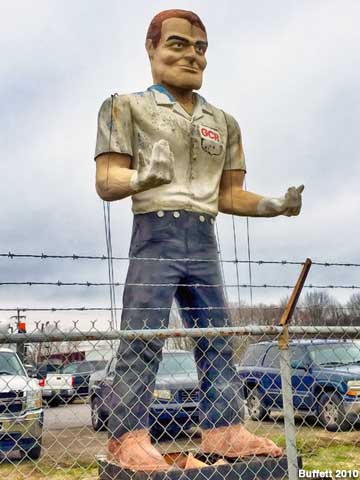 Outdoor full-color statue of a giant man behind a fence wearing a moss-stained white shirt and black pants. His upturned hands are wearing white gloves.