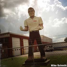 Outdoor full-color statue of a giant man behind a fence wearing a white shirt and black pants. His upturned hands are wearing white gloves.