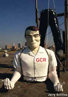 Outdoor full-color statue of a giant man wearing a white shirt and black pants. His upper body sits on the ground, waiting to be attached to his upright legs.