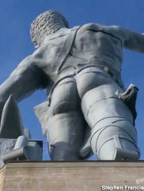 Looking up at the buttocks of a statue of large naked man wearing a leather apron.