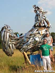 A large, outdoor sculpture of a rooster made with chrome car bumpers. Two people are posed next to it for scale.