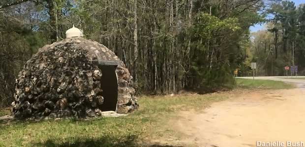 Small, dome-shaped building made of dark rocks has an open doorway, is backed by trees, stands on the edge of a dirt pull-off next to a road.