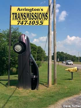 Beneath an outdoor sign for a transmission shop is a 1990s sedan pointing skyward, with its trunk apparently buried in the ground.