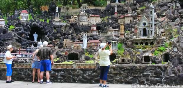 Tourists look up at a rocky hillside filled with miniature models of Mediterranean-style buildings and temples.