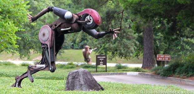 Outdoor sculpture of a sprinter made of metal junk has a motorcycle wheel for a head.