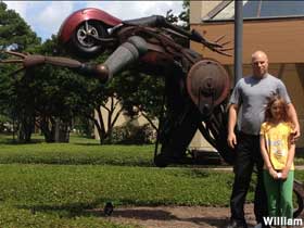 Father and daughter standing in front of an outdoor sculpture of a sprinter made of metal junk with a motorcycle wheel for a head.