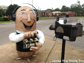 Large outdoor upright fiberglass peanut painted to resemble a man holding a clapboard, wearing headphones, standing in front of a video camera.