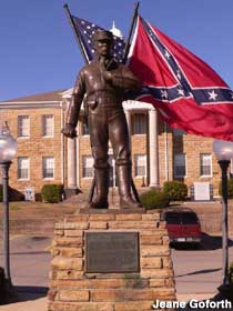 Outdoor bronze soldier stands atop a brick pedestal in front of a USA and Confederate flag.