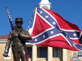 Outdoor bronze soldier stands in front of a USA and Confederate flag.