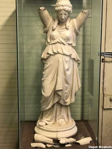 Armless Greek lady statue in a glass case, looks down at her shattered arms and hands at her feet.