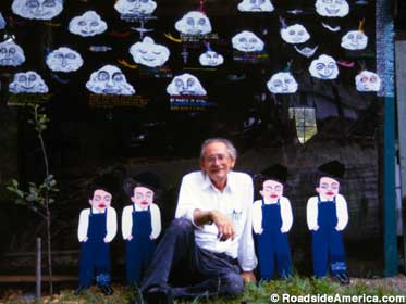 Howard Finster, age 72, sits on the grass in front of his folk art of painted clouds and people.