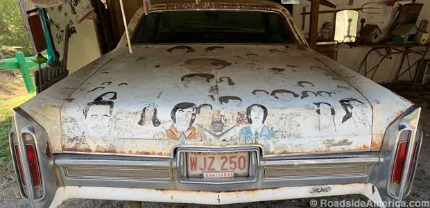 Back end of a 1983 white Cadillac has its trunk lid covered with folk art portraits of faded people.