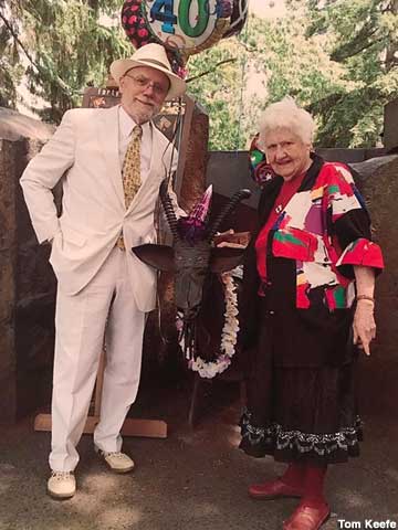 Older man in a white suit and a very old woman wearing a colorful blouse stand on either side of a metal goat wearing a Hawaiian lei and a purple birthday hat.