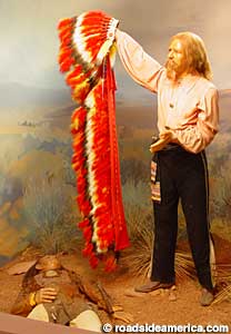 Wax figure of Buffalo bill holds Indian headdress over dead Indian.